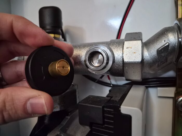 En hand håller i en svart och mässingsfärgad anliggningstermometer framför ett metallrör i en fördelningsinstallation.