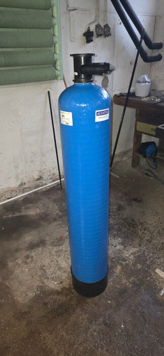 Blå filterbehållare i plast placerad i ett garage med betonggolv och verktygshylla i bakgrunden.