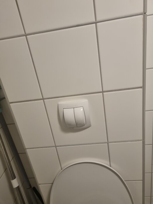 Vitt kakelvägg med spolknapp monterad ovanför en toalettsits, visar tjänstefack för vattenavstängning.