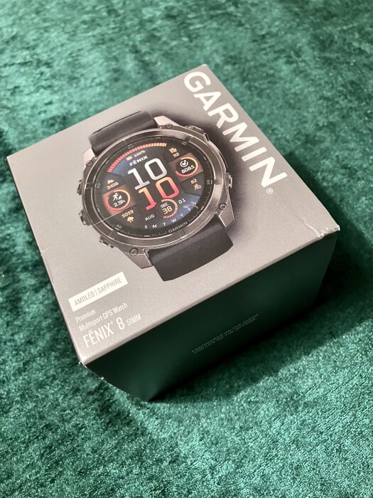Förpackning av Garmin Fenix 8 multisportklocka med AMOLED-skärm, på en grön yta.