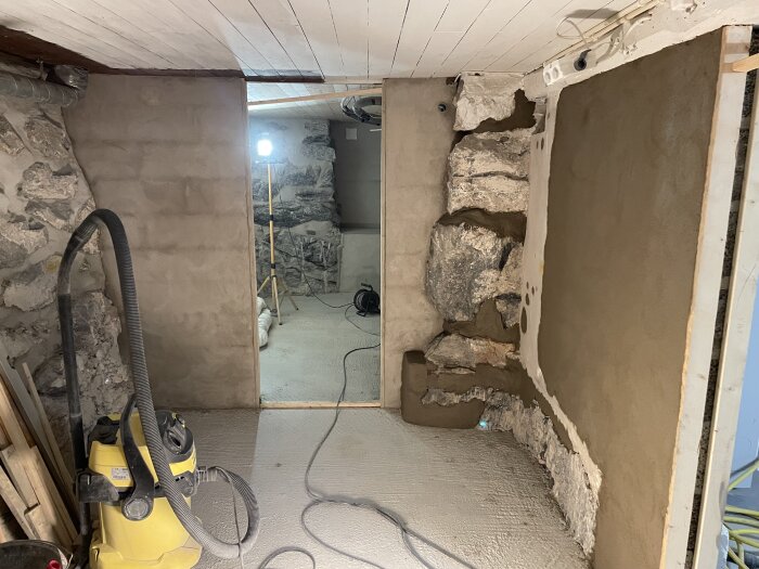 Renoverad källarvägg med puts över sten, en bygglampa i bakgrunden och en våtdammsugare i rummet.