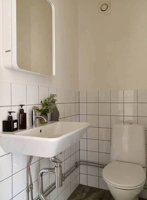 Vitt handfat i modern stil med kromad kran, placerat mot vit kaklad vägg. Tvålpumpar och växt står på bänken. Toilett intill handfatet.