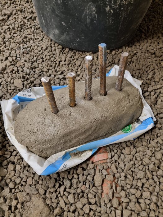 Cementblock med fyra vertikala järnstänger, placerat på en grusig yta, med en plastögla och en svart hink delvis synlig i bakgrunden.