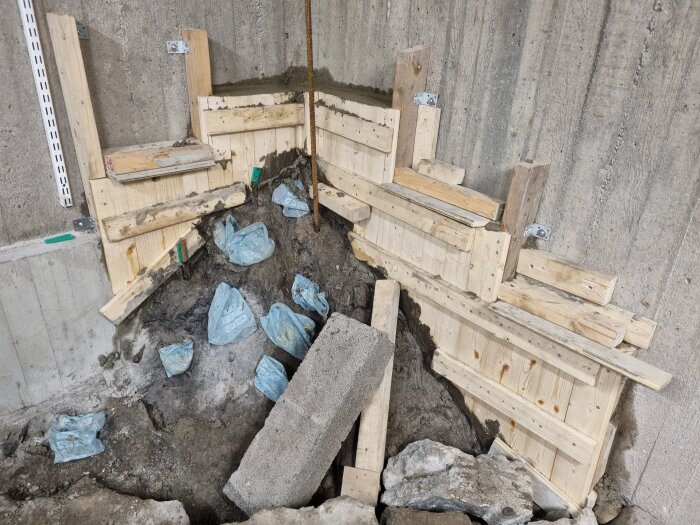 Ett delvis uppbyggt byggprojekt med träformer mot en betongvägg, inklusive några plastpåsar och stenar på marken.