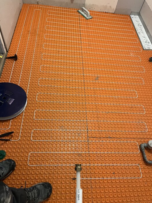 Golvvärmesystem under installation med orange membran och vita rör, verktyg synliga på golvet.