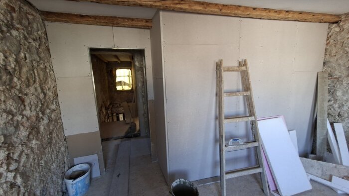 Rum under renovering med stenväggar, nybyggda gipsskivor och en trästege i mitten. Utsikt mot ett rum med fönster i bakgrunden.
