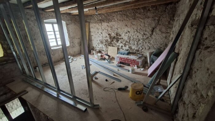 Renovering av ett rum med stående regelverk, oisolerade väggar av sten och ett gammalt fönster. Byggmaterial och verktyg på golvet.