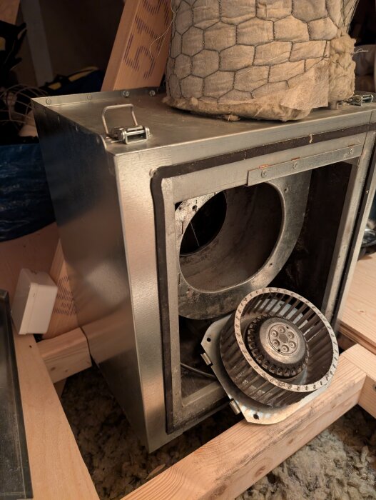 Ventilationsfläkt i metall med demonterad fläkthjul på vinden monterad mellan träreglar, insvept i isolering med hönsnät och akustisk dämpning.