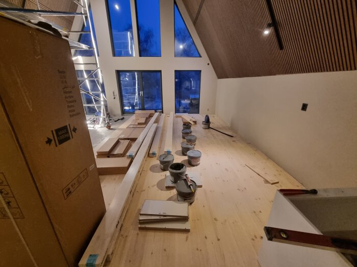 Byggyta med nylagt trägolv, burkar med byggmaterial och delar för köksmontering, stora fönster och högt i tak.