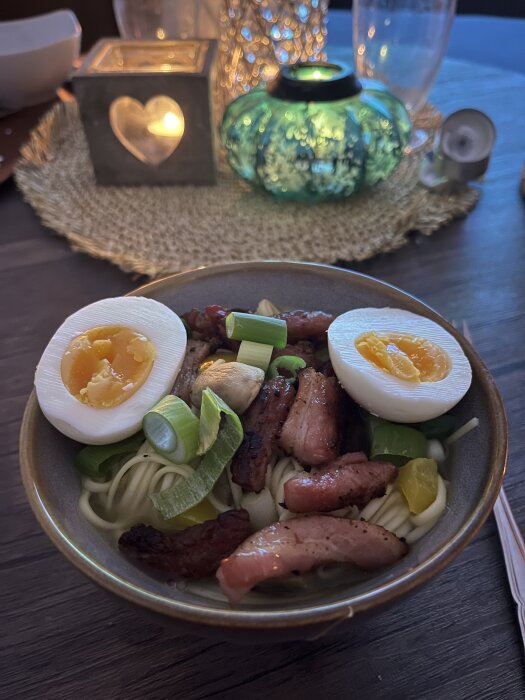 Ramen med stekfläsk, champinjoner, böngroddar, salladslök, paprika, nudlar och kokt ägg i en skål på dukat bord med tända ljus.