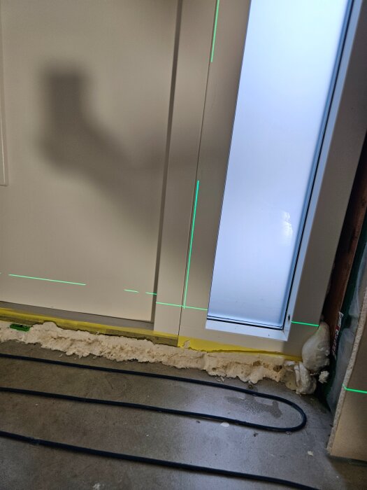 Byggnadsarbete med dörrkarm, synlig fogmassa och golvvärmeslangar; gröna laserlinjer markerar nivåer.