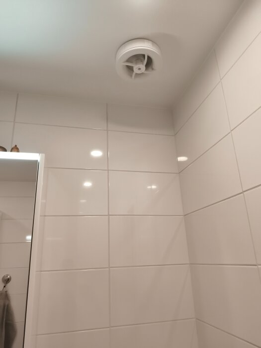 Helrenoverat badrum med vitkaklade väggar och en takventil synlig i hörnet, reflekterande ljus från spotlights i taket.