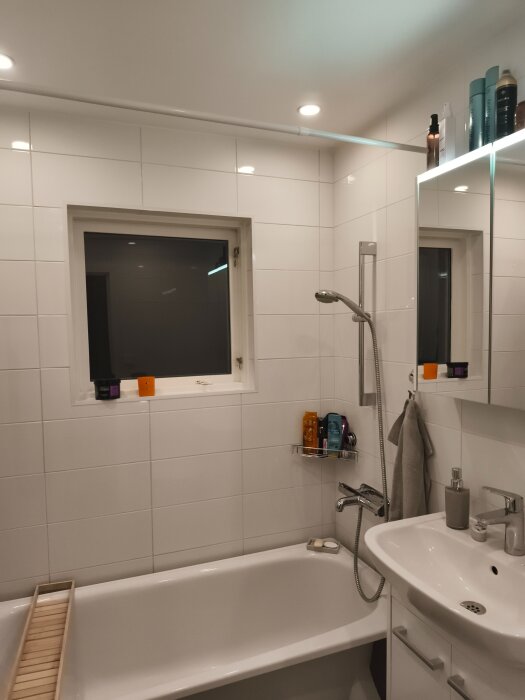 Renoverat badrum med vitt kakel, badkar, handfat och duschhandtag. Rengöringsprodukter på hylla och fönsterbräda, modern och fräsch stil.