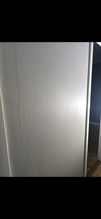 Vägg med ojämn målning och spackling, visar textur och skuggor där målningen inte är slät, ljus belysning, del av dörrkarm synlig.