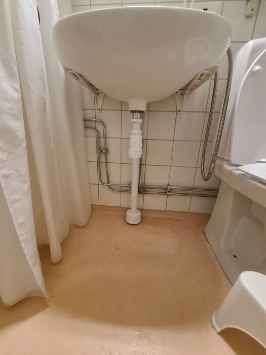 Handfat med synlig rördragning under, duschdraperi till vänster och toalett till höger, på beige badrumsgolv.