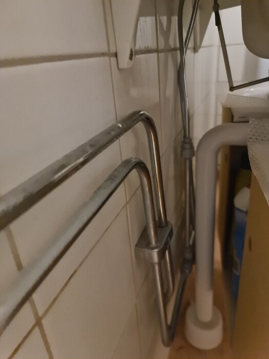 Rörsystem under handfatet, inklusive två horisontella metallrör och ett vitt avloppsrör, monterade mot en kaklad vägg.