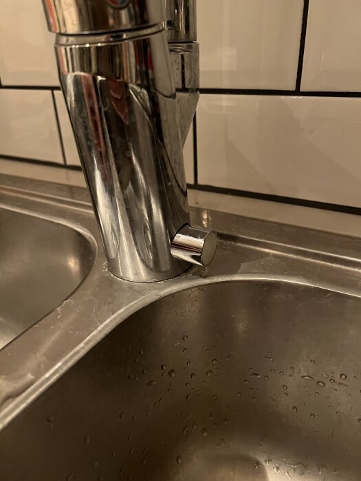 Rostfri kökskran med knapp på diskbänken, bredvid diskho med vattenstänk. Problem med glappande elknapp kopplat till diskmaskinen.