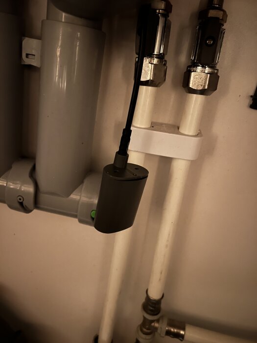 Grå plasthylsa med grön knapp ansluten till rör och slangar under diskbänk. Elektrisk installation för diskmaskinens vattenflöde.