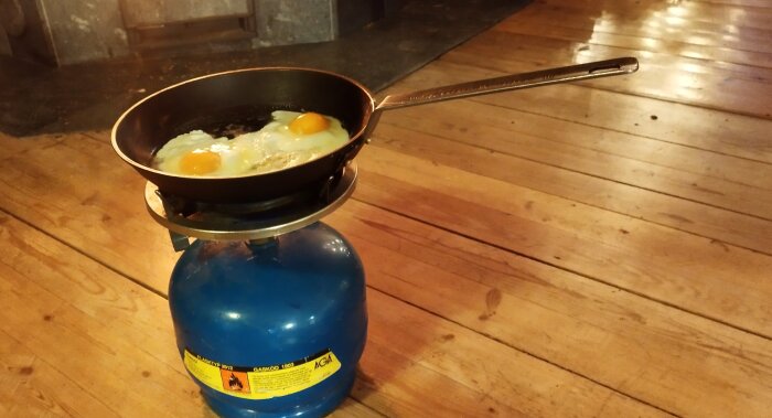 Stekpanna med stekta ägg på en gasolbrännare i ett rum med trägolv, använd för matlagning vid strömavbrott.