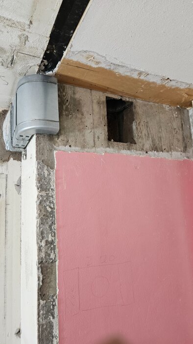 Rektangulär ventilationskanal monterad i hörnet av ett rum med en rosa vägg, omgiven av rå betong och gips.