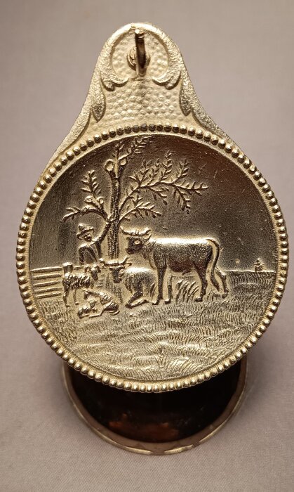 Guldfärgat ställ för fickur, med relief av bondgårdsscen, träd och betande kor. Märkt med Tyska rikets patent.