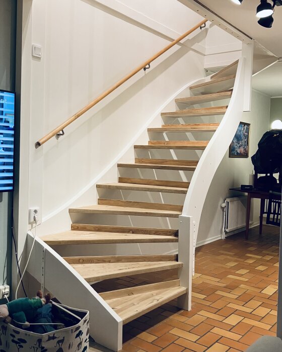 En renoverad trätrappa med ljus målning och en svängd design, kompletterad med ett enkelt trähandtag, mot en vägg i ett rum med kakelgolv.