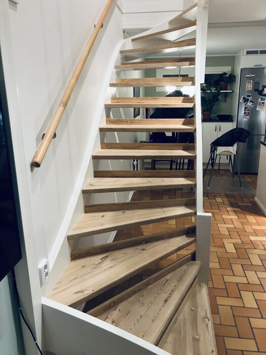 Renoverad trappa med ljusa trappsteg i trä och ett handräcke, omgiven av ljusa väggar och ett modernt kök i bakgrunden.