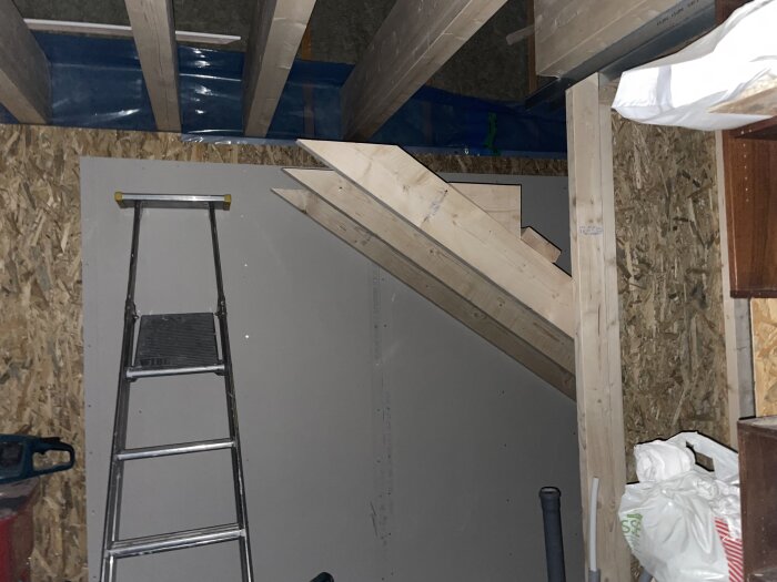 Byggarbetsplats med träbjälkar och en stege lutad mot en vägg av spånskivor, pågående konstruktion inomhus.