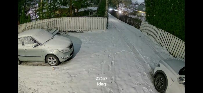 Snöig uppfart med två bilar, sett genom kamera; vintermiljö med träd och stängsel; inspelningstiden visar 22:57.