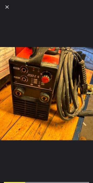 Röd och svart pinnsvetsmaskin modell 200 E 5, stående på ett träbord med sladdar och kontrollpanel synliga.