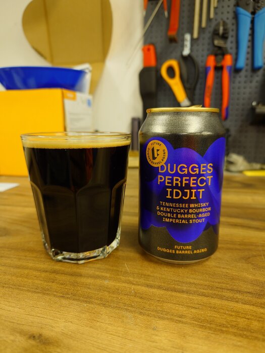 En öl från Dugges, "Perfect Idjit", med en fylld glas bredvid, i en verkstadsmiljö med verktyg i bakgrunden.