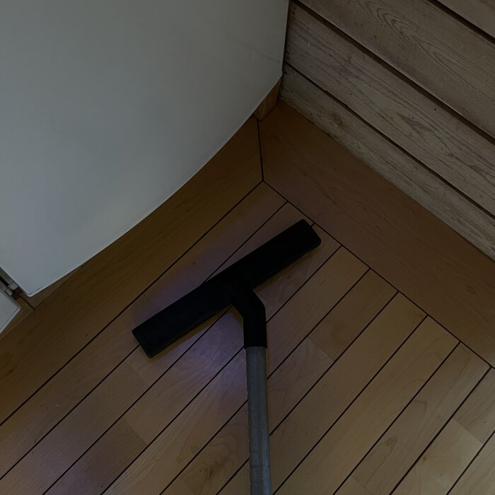 Dammsugarmunstycke med lång slang på en trägolv nära en vägg.