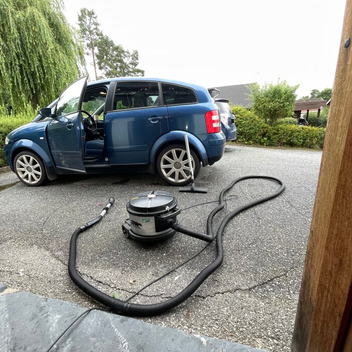 Centraldammsugare på uppfart bredvid en blå bil med öppen dörr och lång slang, perfekt för rengöring av bilen.