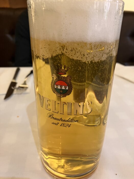 Ett glas med skummande öl av märket Veltins på ett dukat bord.