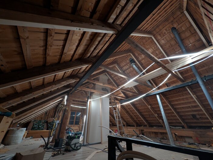 Renovering pågående i ett timmerhus, synligt tak med takbjälkar och installationer. Ljusinsläpp från takfönster och byggmaterial på golvet.