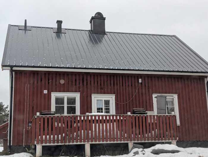 Rött timmerhus med svart pannplåtstak, snötäckt mark och veranda. Två skorstenar ses på taket.