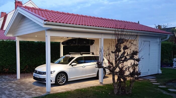 Vit carport med rött tak och smala trästolpar, en vit bil parkerad under, intilliggande förråd med grå dörr, omgiven av stenlagd mark och grönska.