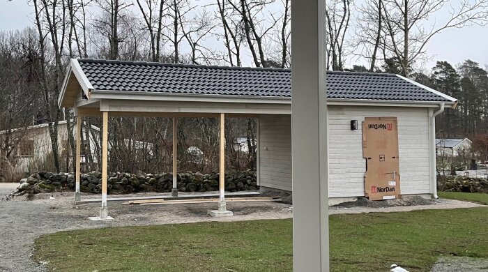Nybyggd carport med förråd, tunna trästolpar under takstommen, förrådsdörr täckt av kartong. Omringad av träd och stenmur i bakgrunden.