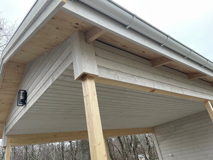Träcarport med ljus träpanel, smala stolpar och en svart vägglampa, omgiven av träd i bakgrunden.