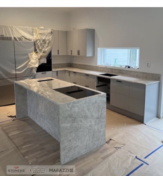 Modernt kök under renovering med grå bänkskivor och plast täckande apparater, vissa skåpluckor saknas.