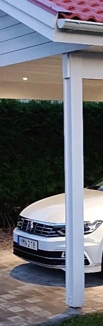 En vit carport med stående stolpar täckta av dekorativa paneler, med en vit bil delvis synlig bakom en stolpe.