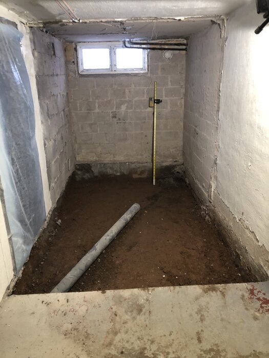 Källare med jordgolv, synliga rör och väggar av blåbetong. En tumstock står mot väggen och ett PVC-rör ligger på marken.