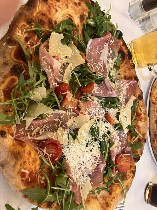 Pizza med parmaskinka, ruccola, tomater och hyvlad parmesan, serverad på en tallrik med ett glas öl i bakgrunden.