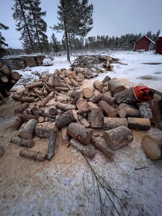 Mängd vedklabbar ligger utspridda i snöigt landskap, redo för klyvning. Bakgrunden visar staplade vedhögar och snöklädda träd.