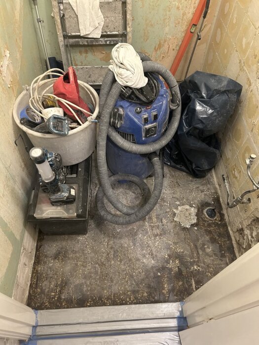 Våtutrymme under renovering med borttaget kakel och synligt vinylgolv; verktyg och dammsugare står på golvet.