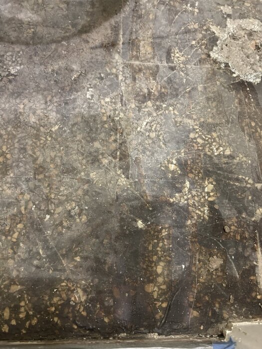 Sliten betongyta med spår av sten och grus, delvis täckt av fläckar och sprickor, vid kanten ett fragment av bortnött material.