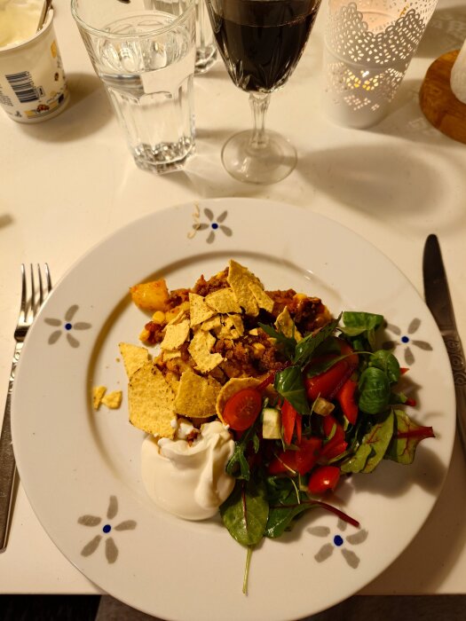 Tacopaj serverad på en tallrik med nachochips, sallad och gräddfil, bredvid ett glas rött vin och ett glas vatten på ett dukat bord.