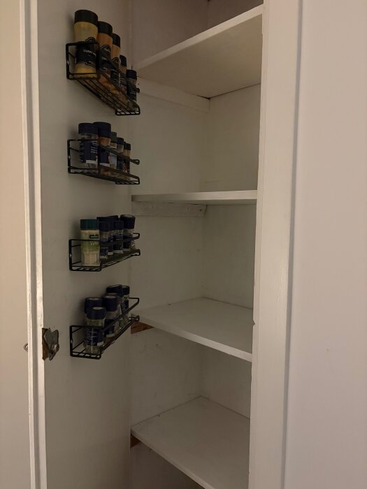 Ett vitt, nybyggt skafferi med hyllor och kryddhyllor på insidan av dörren, nästan färdigställt.