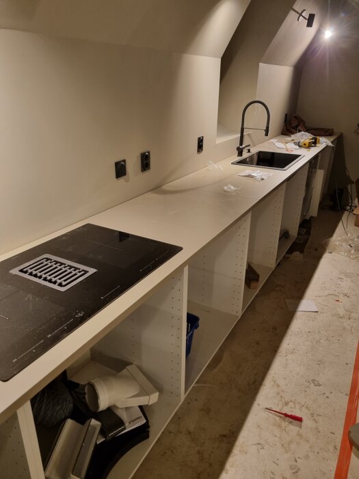 Köksinstallation med spishäll och diskbänk under arbete i en halvfärdig köksenhet, med belysning ovanför arbetsytan.