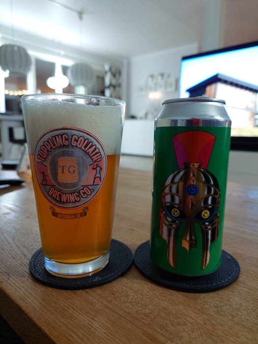 Ett fyllt ölglas med Toppling Goliath-logotyp bredvid en färgglad ölburk med hjälmmotiv på ett bord.
